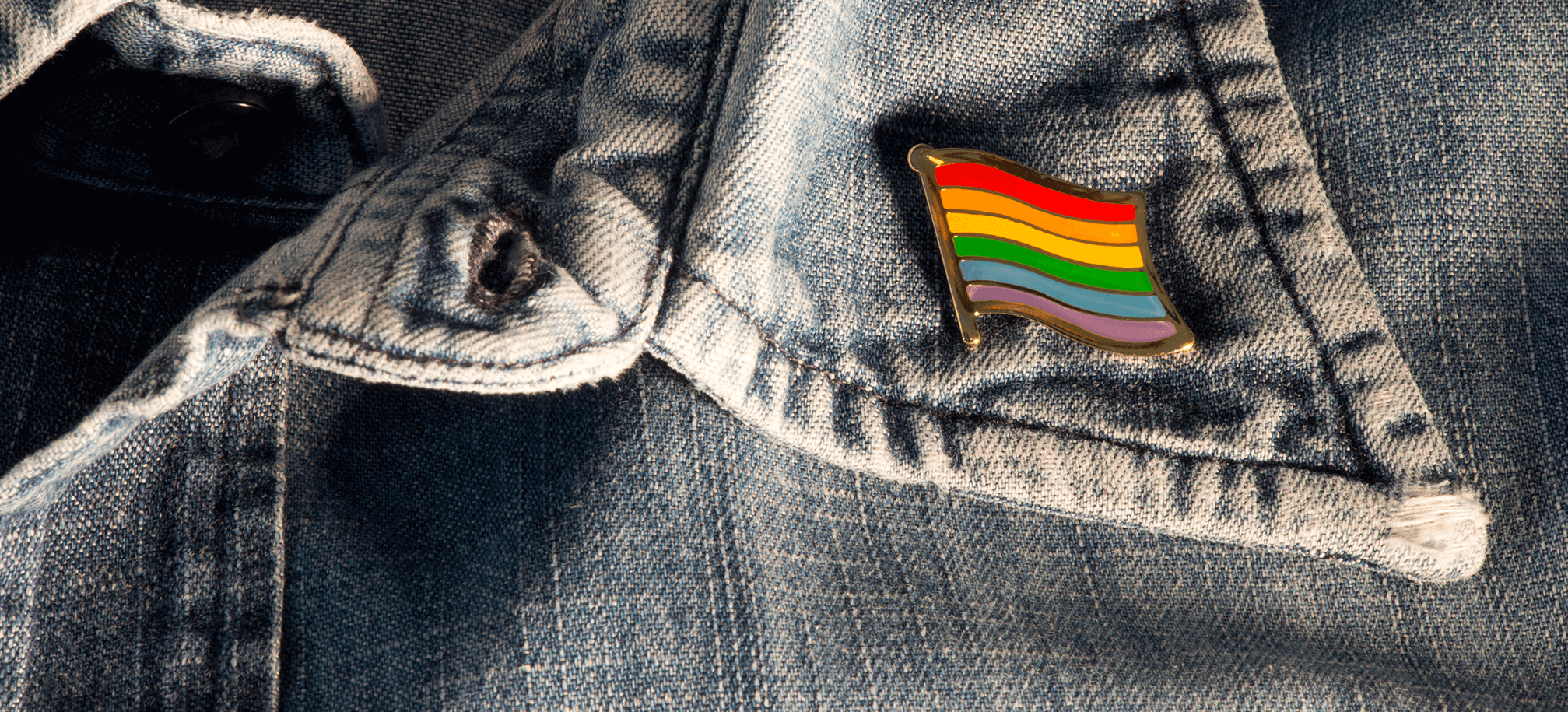 Close up image of a denim jacket with a pride flagged pinned to the collar